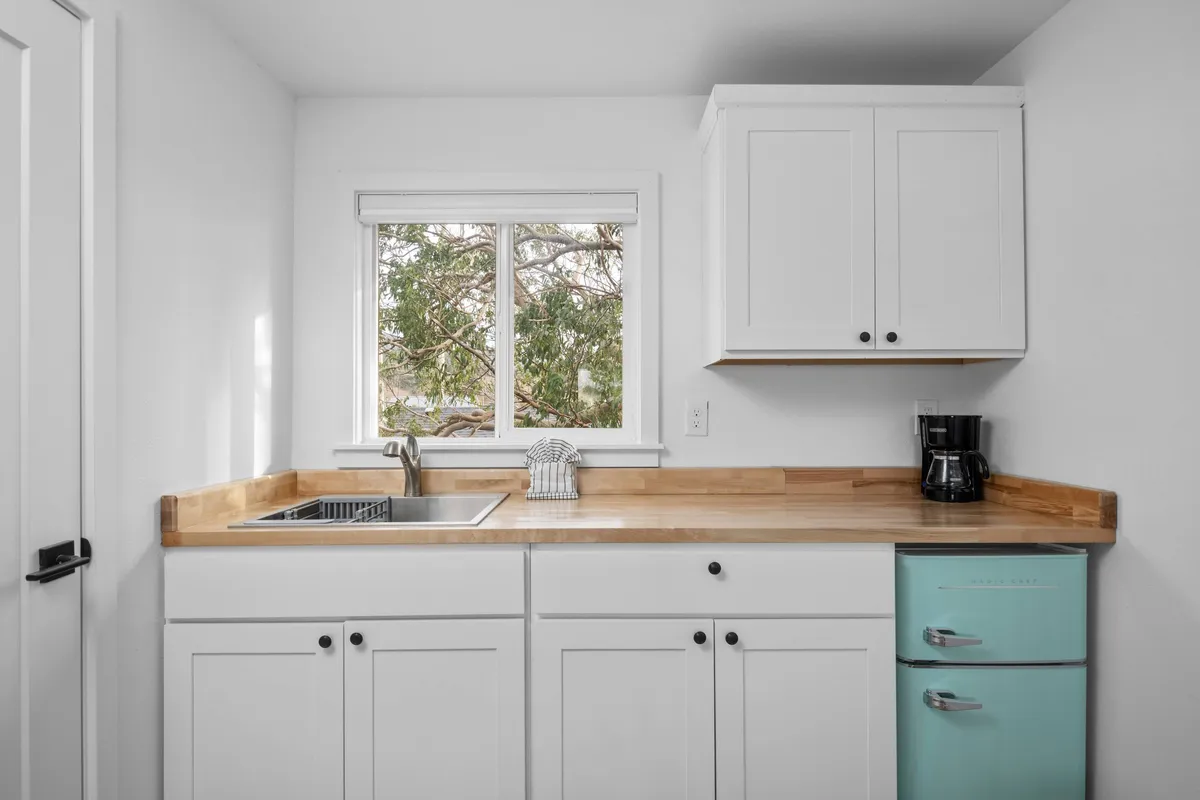 This image shows a cozy kitchenette featuring a wooden countertop, a mint green refrigerator, a coffee maker, and a window with a view of trees. The countertop is clear and ready for meal prep, and the space feels bright and inviting, ideal for enjoying a relaxed morning coffee or preparing light snacks.