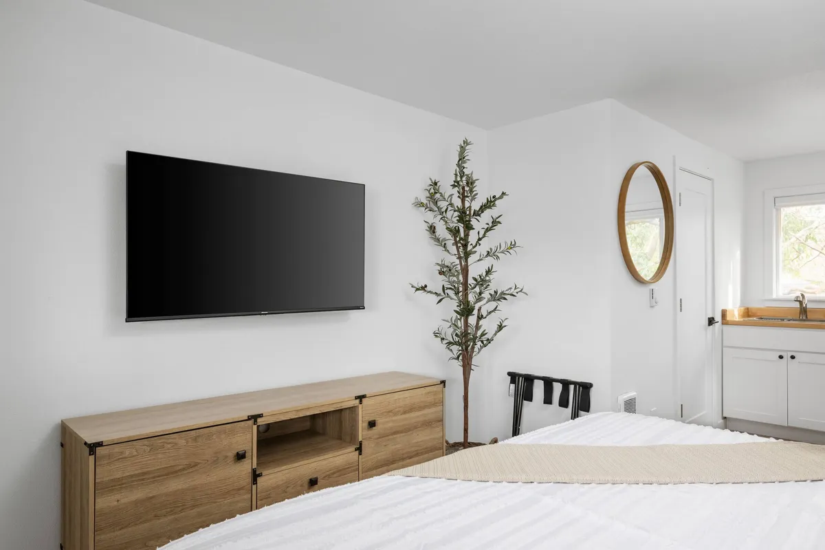 The image showcases a tranquil condo living area featuring a comfortable seating arrangement facing a television, warm wooden furniture, modern mirrored round wall decor, and a touch of greenery from an indoor plant. The inviting space feels freshly updated, perfect for unwinding with a favorite show or a good book, with natural light enhancing the calm ambiance.