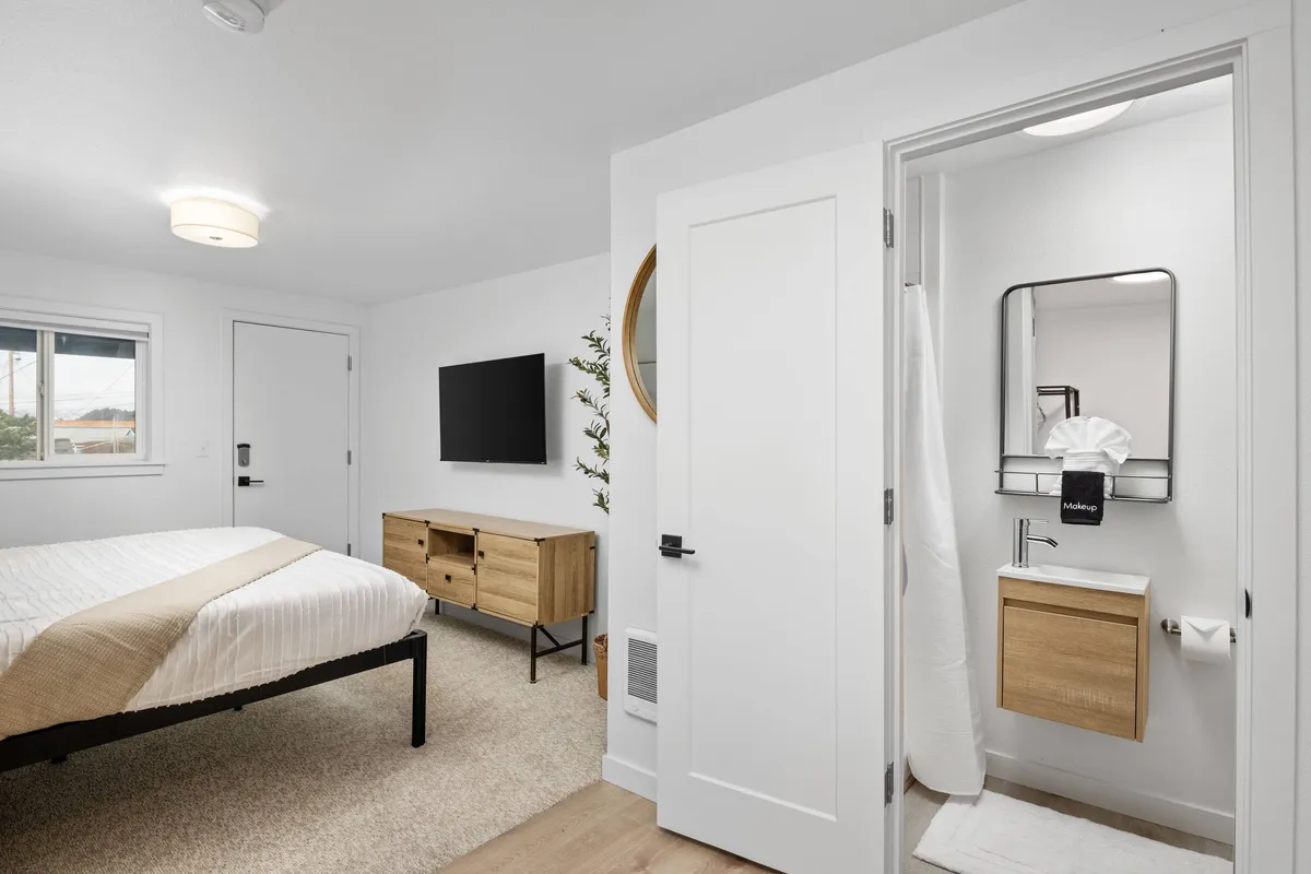 This image provides a bright view of the bedroom, showcasing a spacious bed with crisp linens, a modern television, dresser, mirror, and the entrance to the bathroom. Soft natural light enhances the fresh, remodeled appeal of the room and highlights its comfortable and modern ambiance.
