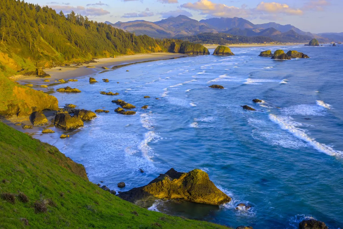 This image captures a wide, sandy stretch of Rockaway Beach bordered by a rocky coastline, with gentle waves rolling in and distant mountains visible on the horizon. The tranquil atmosphere is perfect for beachcombing, photography, or simply enjoying the natural beauty of the Oregon coast, especially as the sun sets over the water.