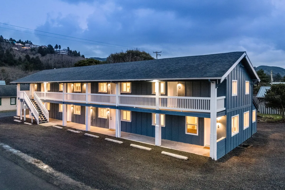 This image showcases the blue exterior of the inn-style building with warmly illuminated windows and a visible exterior staircase. There are guests socializing on the balcony, suggesting a lively, communal atmosphere. The inviting glow from the windows hints at the privacy and comfort available inside each unit, while the balcony and staircase highlight opportunities for spontaneous guest interactions and easy movement between spaces.